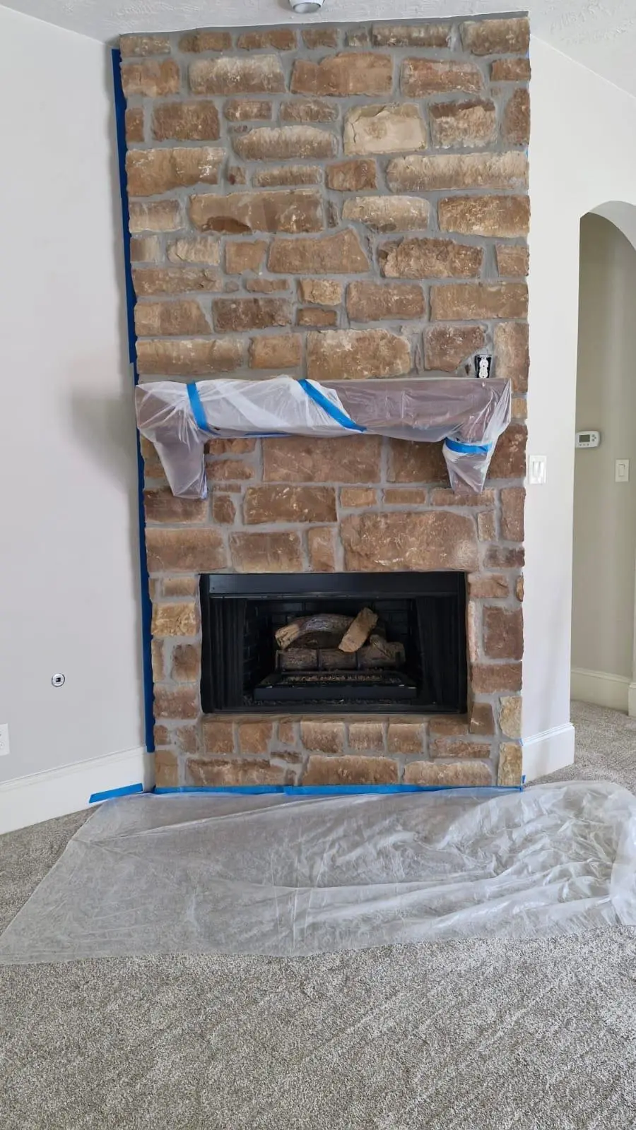 Stone fireplace before painting, with the mantel wrapped in plastic, blue tape on the walls, and plastic covering the carpet